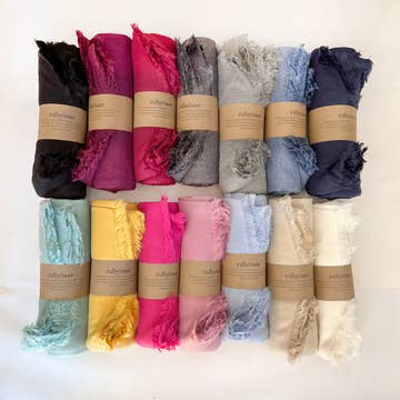 Twelve Hima Large Scarves—cashmere, fringed, in black, dark pink, magenta, grey, light blue, navy, mint, yellow, bright pink, light pink, pale blue,and ivory—rolled with a brown label. Fair trade scarves are arranged in three rows on a white background.
