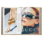 An open copy of the *Little Book of Gucci* features an advertisement on its pages. On the right, a woman wearing oversized sunglasses looks upward, epitomizing Italian fashion elegance. The left side showcases a model's legs adorned with gold high-heeled sandals, holding a fringed gold handbag. The luxury brand name "GUCCI" is prominently displayed.