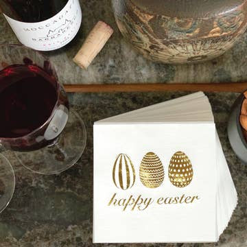 A stack of holiday-themed cloth-like cocktail napkins, featuring "happy easter" and gold-patterned eggs, sits on a marble surface. Nearby are hosting accessories: a filled wine glass, an uncorked wine bottle with its cork, a decorative container, and a bowl of nuts.