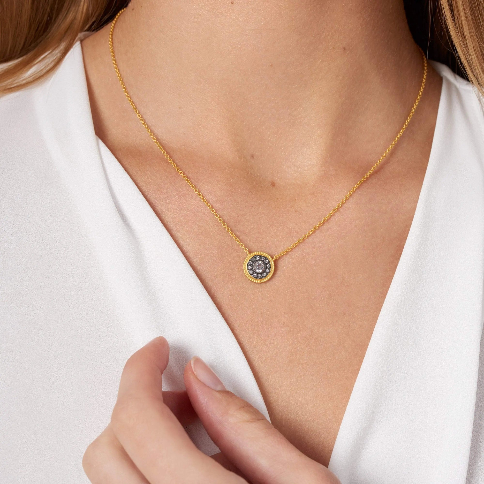 A woman in a white blouse models the Nautical Button Necklace, featuring a round 14K gold pendant with a dark central stone, surrounded by cubic zirconia. Her hand gently touches her blouse near the necklace, emphasizing its elegance against her skin.