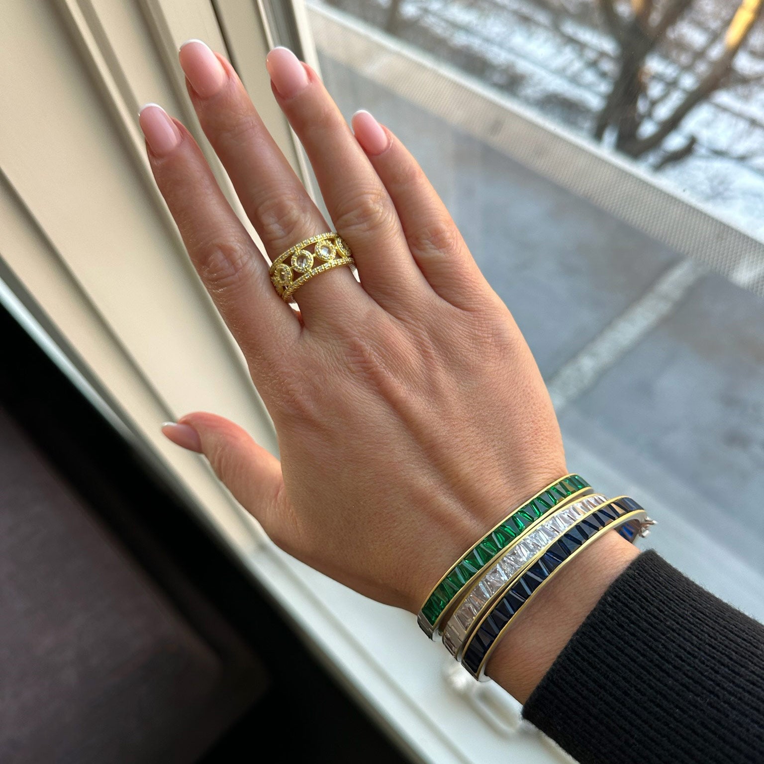 A hand with light pink nails flaunts a Kaleidoscope Cutout Cigar Band Ring on the middle finger, alongside green, blue, and silver bracelets. A snow-dusted street can be seen through the window, and a black sleeve is partially visible.