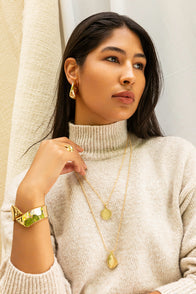 A woman with long dark hair wears a cream turtleneck sweater, gold earrings, layered necklaces, and the Alexandra Overlap Cuff. She gazes to the side, resting her hand near her chin against a backdrop of beige fabric textures.