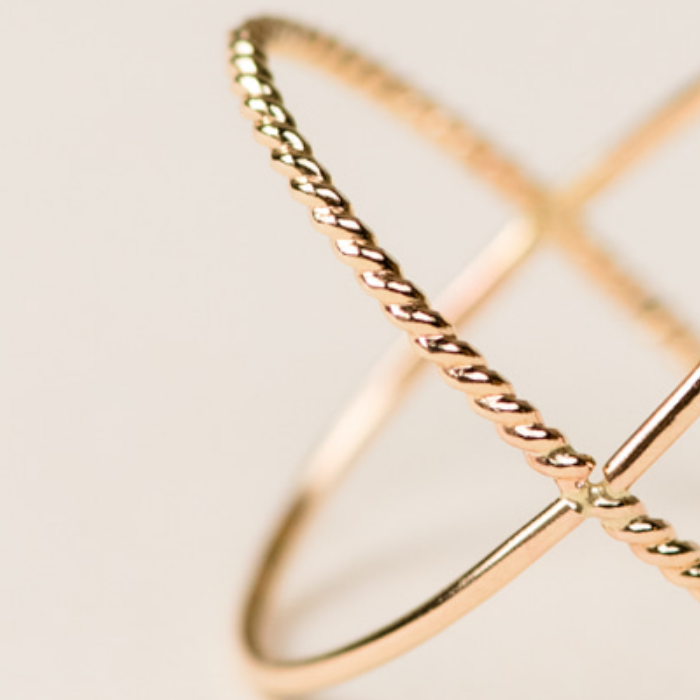 Close-up of a delicate Braided X Ring on a soft beige background. This 14k gold-filled handcrafted ring features a minimalistic design with twisted detailing and overlapping bands, creating an elegant, interconnected pattern. The lighting highlights the shiny, polished texture of the metal.