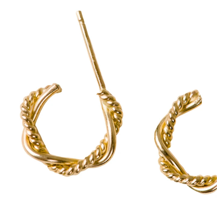 A close-up of a 14k gold-filled Braided Mini Hoop earring, featuring a rope-like texture, rests on a flat, off-white surface. The earring's post extends upwards while another matching piece is partially visible, indicating it's part of the Braided Mini Hoops pair. The lighting accentuates the shiny and intricate detailing of these everyday earrings.
