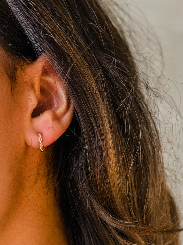 A close-up captures a person's ear adorned with Braided Mini Hoops crafted from 14k gold-filled material. The individual's long, brown hair cascades softly, partially hiding the ear. The blurred and neutral background accentuates the earring's understated elegance and highlights the luxuriant texture of the hair.