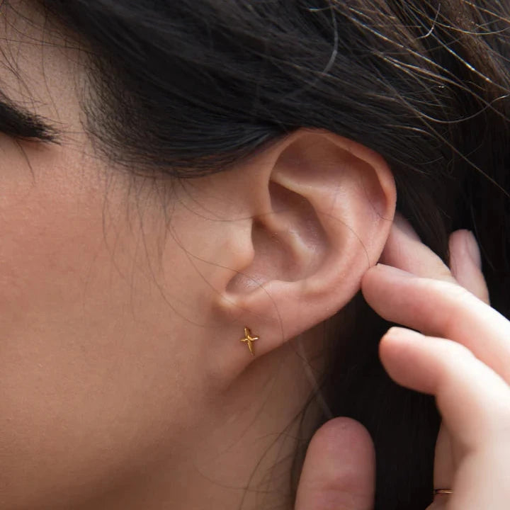 Close-up of a person's ear adorned with the Droplet Cross Studs, part of an elegant earring stack. The dark hair is tucked behind the ear, and they gently touch the back of the ear with their fingers. The skin is smooth, and the earrings subtly catch the light, highlighting their simplicity and elegance.