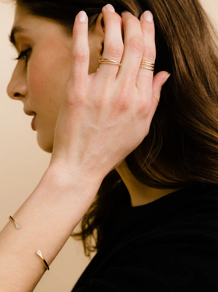 A woman with long brown hair is touching her head, showcasing several delicate gold-fill pieces from the Virgil Ring collection. She also wears a slim gold bracelet on her wrist. Set against a soft beige background, she is dressed in a black top, and her short nails complement the elegance of her handmade jewelry ensemble.