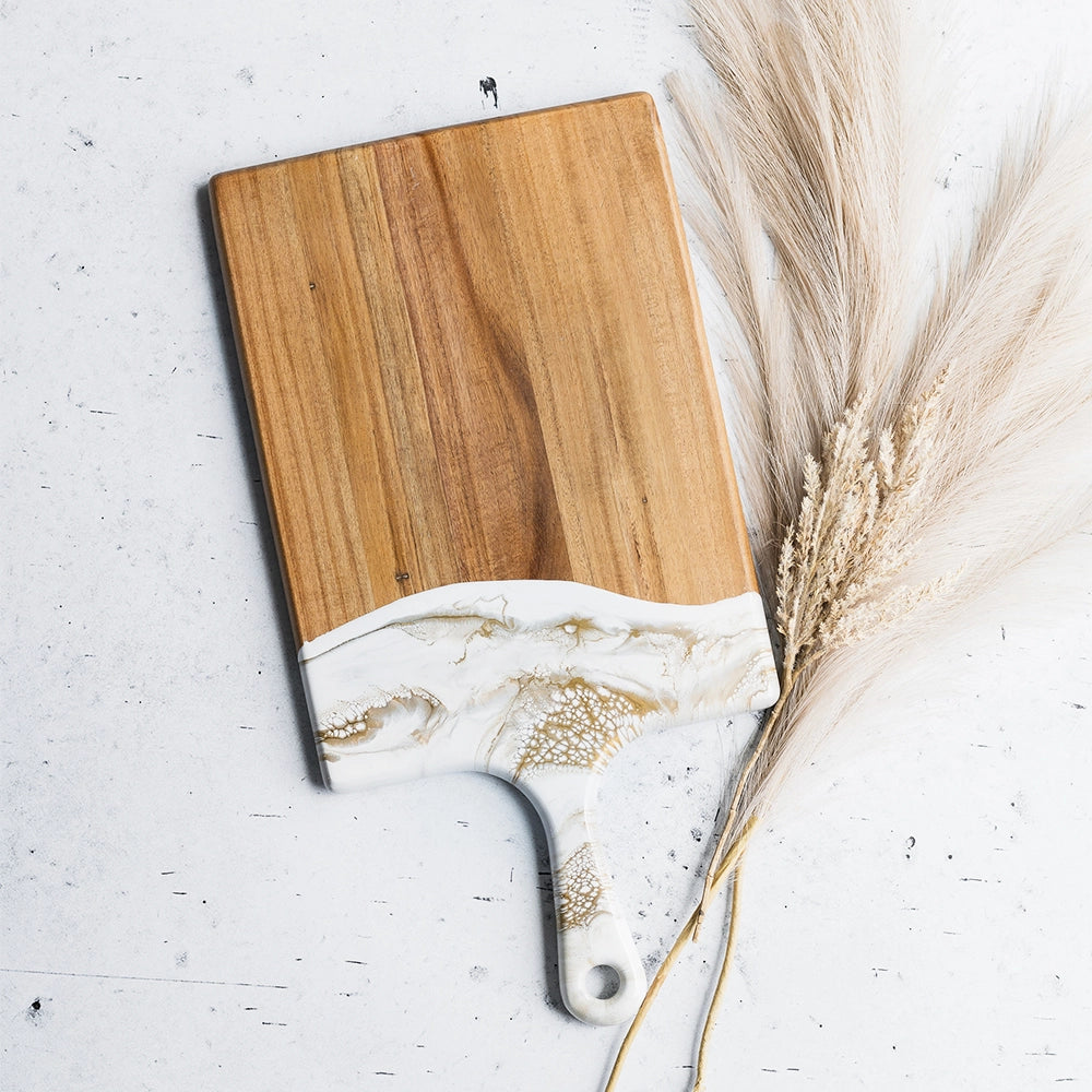 The Resin Acacia Cheese Board features a glossy white handle with gold accents and is crafted from eco-friendly resin. Its rectangular shape with a curved bottom edge rests on a textured white surface, complemented by elegant beige pampas grass stalks nearby.