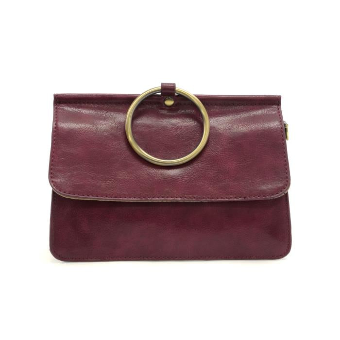 The Aria Ring Bag - Fall/Winter is a burgundy leather clutch featuring a fold-over flap and a prominent gold circular handle, giving it the appearance of a bracelet bag. It has a smooth texture and showcases a minimalist design, with neatly stitched edges that highlight its clean lines and elegant style.
