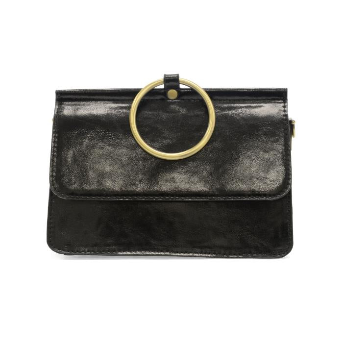 The Aria Ring Bag - Fall/Winter is a sleek black leather clutch with a glossy finish, highlighted by a large gold ring serving as the top handle above the flap closure. This minimalist bracelet bag features the ring as its distinctive focal point, and its texture is smooth and polished.