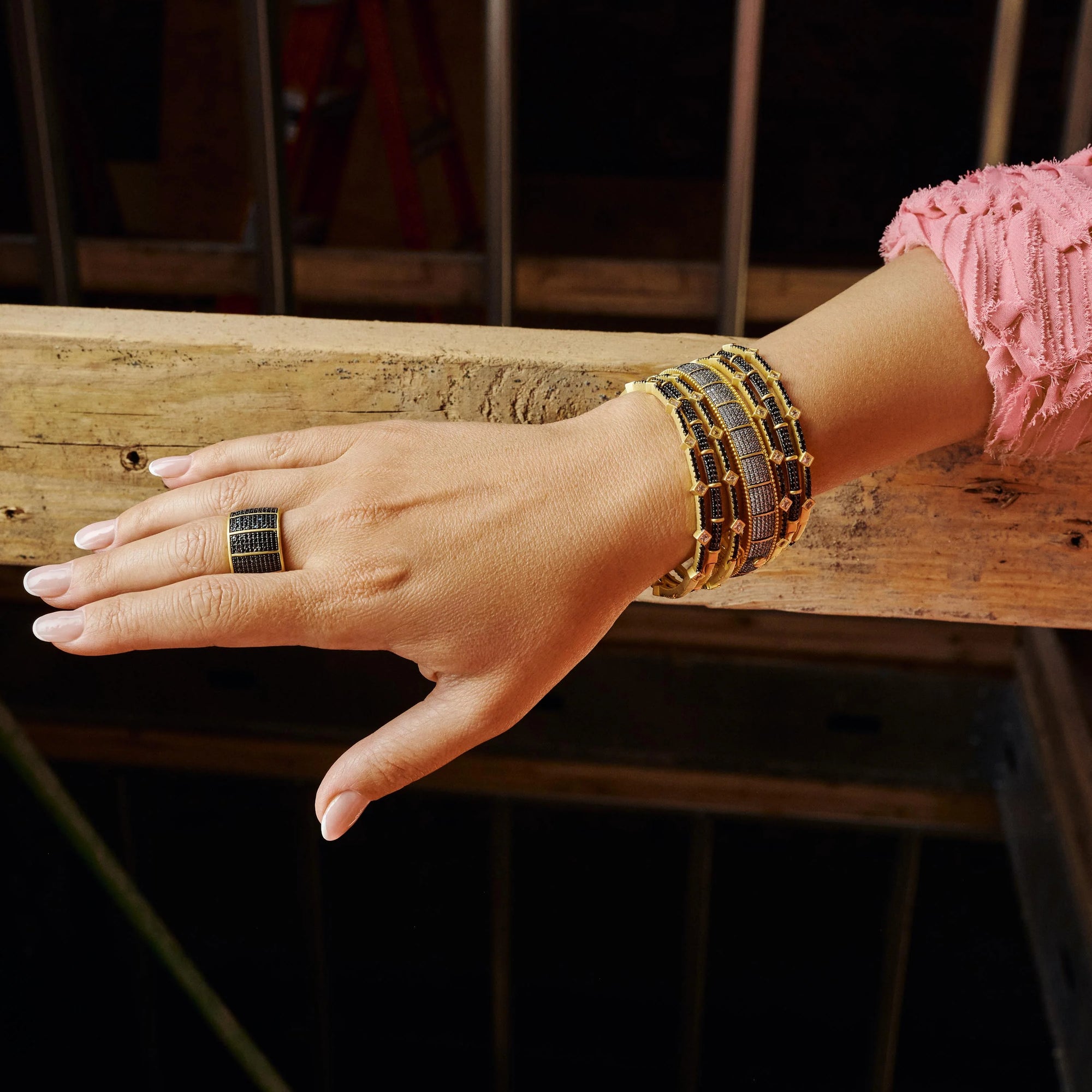 A hand with light skin, styled with a black and gold ring and several matching beaded bracelets, rests on a rustic wooden railing. The person is wearing a textured pink sleeve and an eye-catching Industrial Finish Cobblestone Statement Hinge Bracelet. The backdrop features blurred vertical metal bars in a dimly lit space.