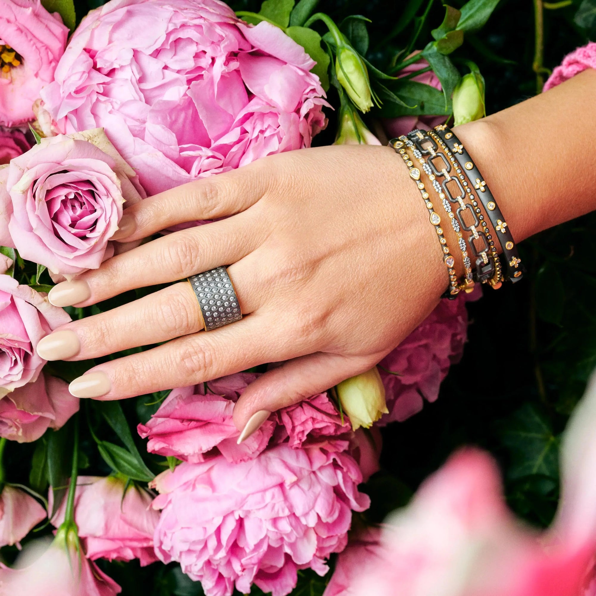 A hand with beige nails is adorned with a silver ring and The New Icon Cigar Band, accompanied by three layered bracelets resting on a cluster of pink and light pink roses. The bracelets are embellished with small gold accents and cubic zirconia stones, adding a touch of elegance to the floral background.