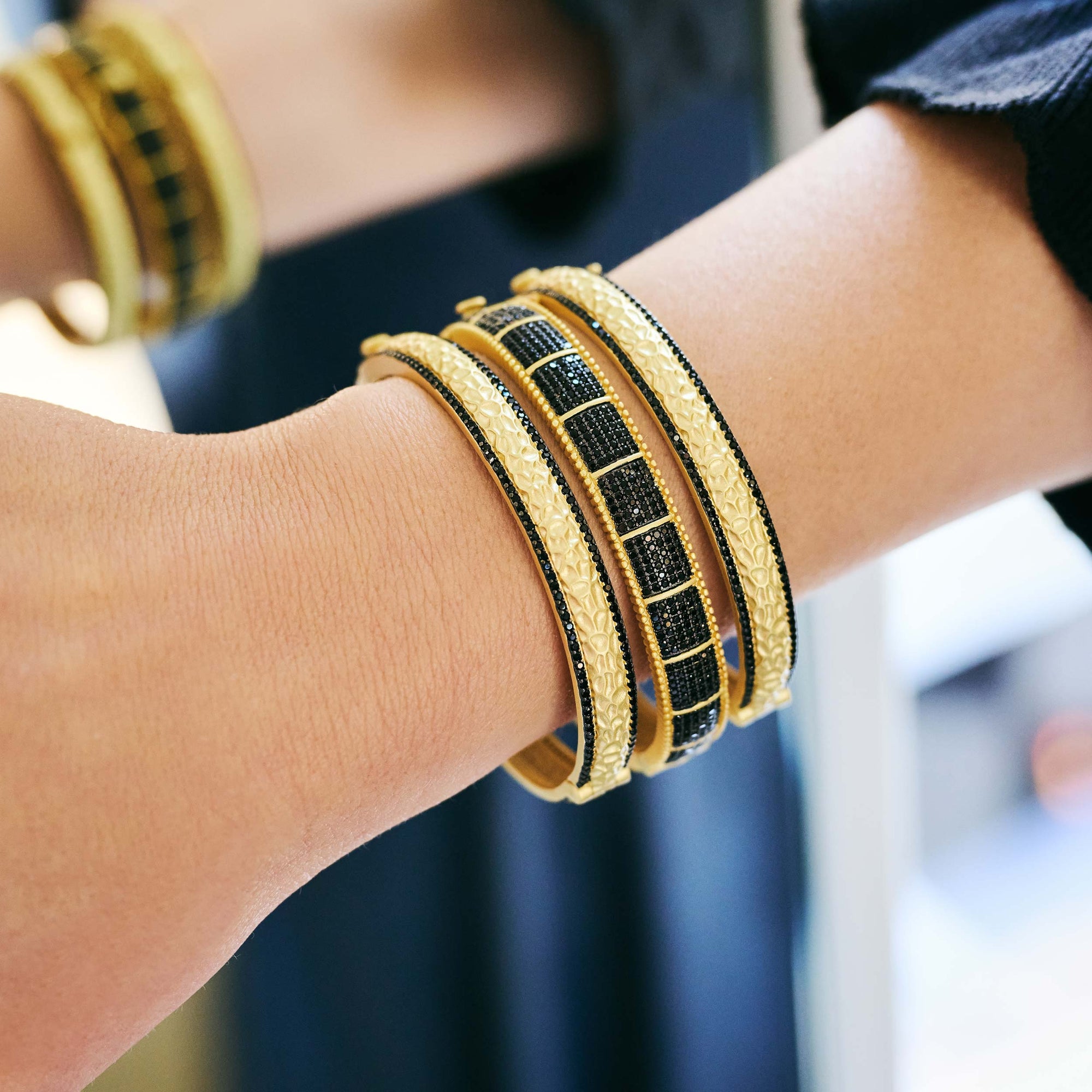 Close-up of a person wearing several elegant bracelets on their wrist, featuring the stunning Industrial Finish Cobblestone Statement Hinge Bracelet. The gold bracelets showcase intricate patterns and are embellished with black accents and cubic zirconia stones, set against a softly blurred background that highlights their luxurious details.