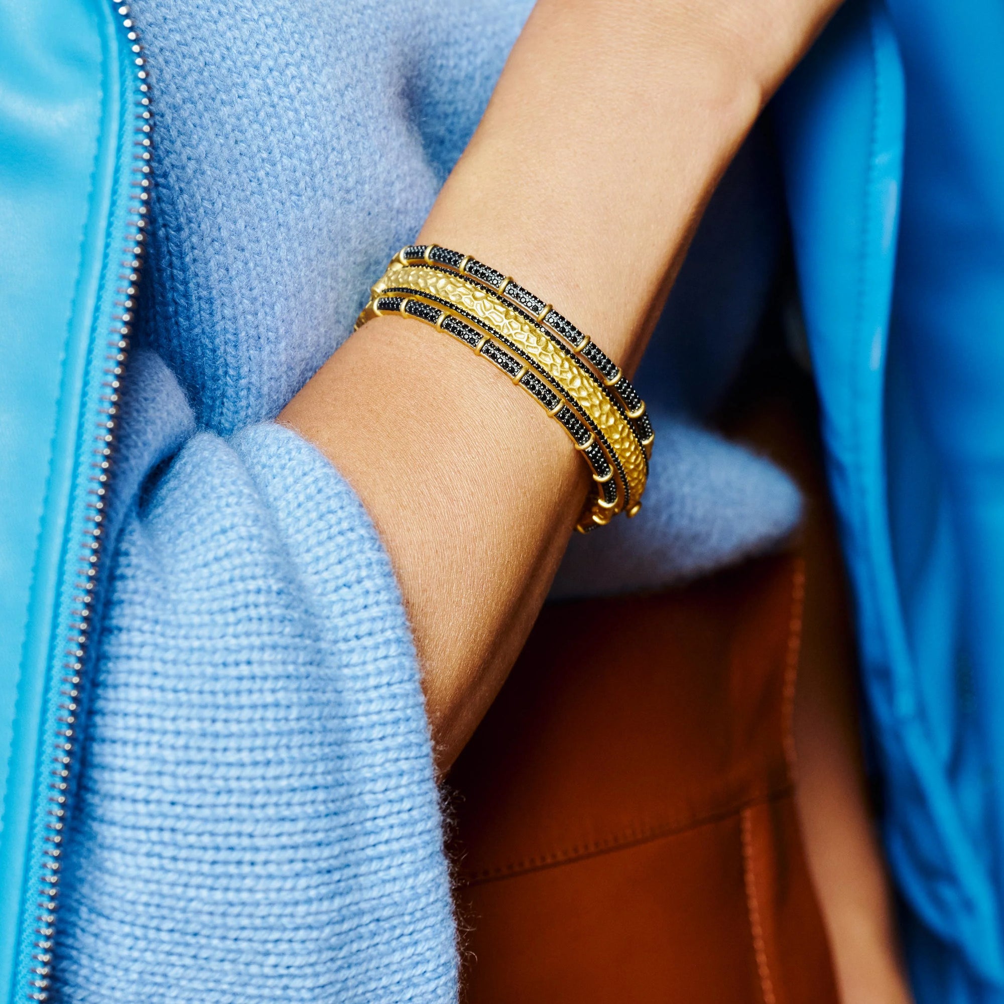 A close-up photo features a person wearing a fluffy blue sweater and jacket, with their hand tucked inside a brown leather bag, adorned by the Industrial Finish Textured Hinge Bracelet embellished with dark stones and cubic zirconia.