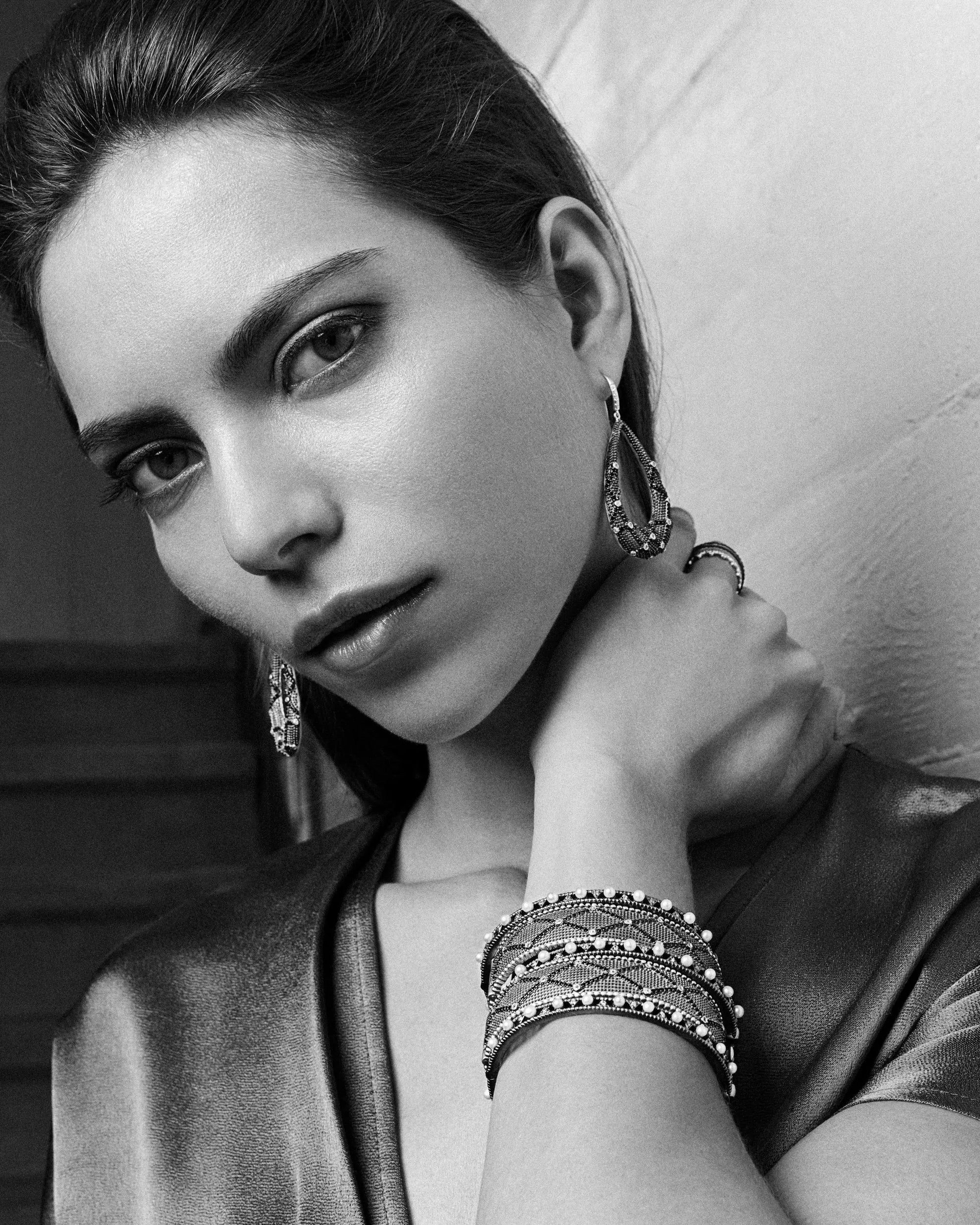 A black and white photograph captures a woman with long hair pulled back, wearing hoop earrings and the Signature Textured Hinged Bangle with a 14K gold finish layered elegantly on her wrist. Her hand gently rests on her neck, complementing her satin-like top. The soft-focus background features stairs, evoking a Brooklyn Bold vibe.