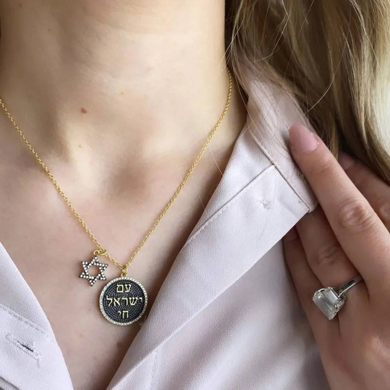 A person wears a light pink blouse paired with the Star of David Pendant Necklace, which symbolizes "Am Yisroel Chai" and features Hebrew lettering. Complementing this is a large, rectangular-cut gemstone ring that adorns the left hand resting on their chest, enhancing the symbolic jewelry ensemble.