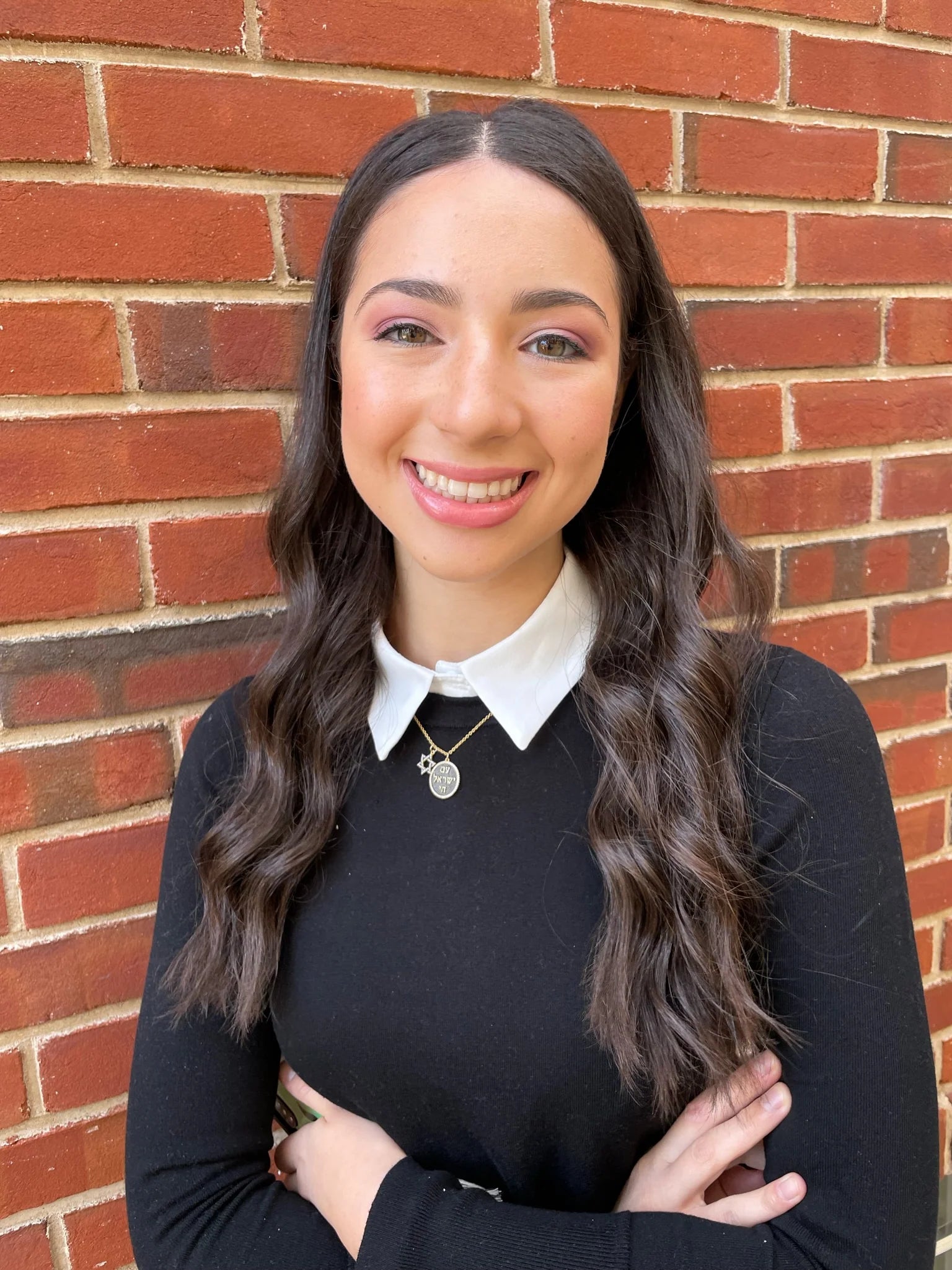 A smiling individual with long, wavy brown hair stands in front of a red brick wall. They wear a black top with a white collar and a Star of David Pendant Necklace. With their arms casually crossed, the natural lighting highlights their friendly expression.