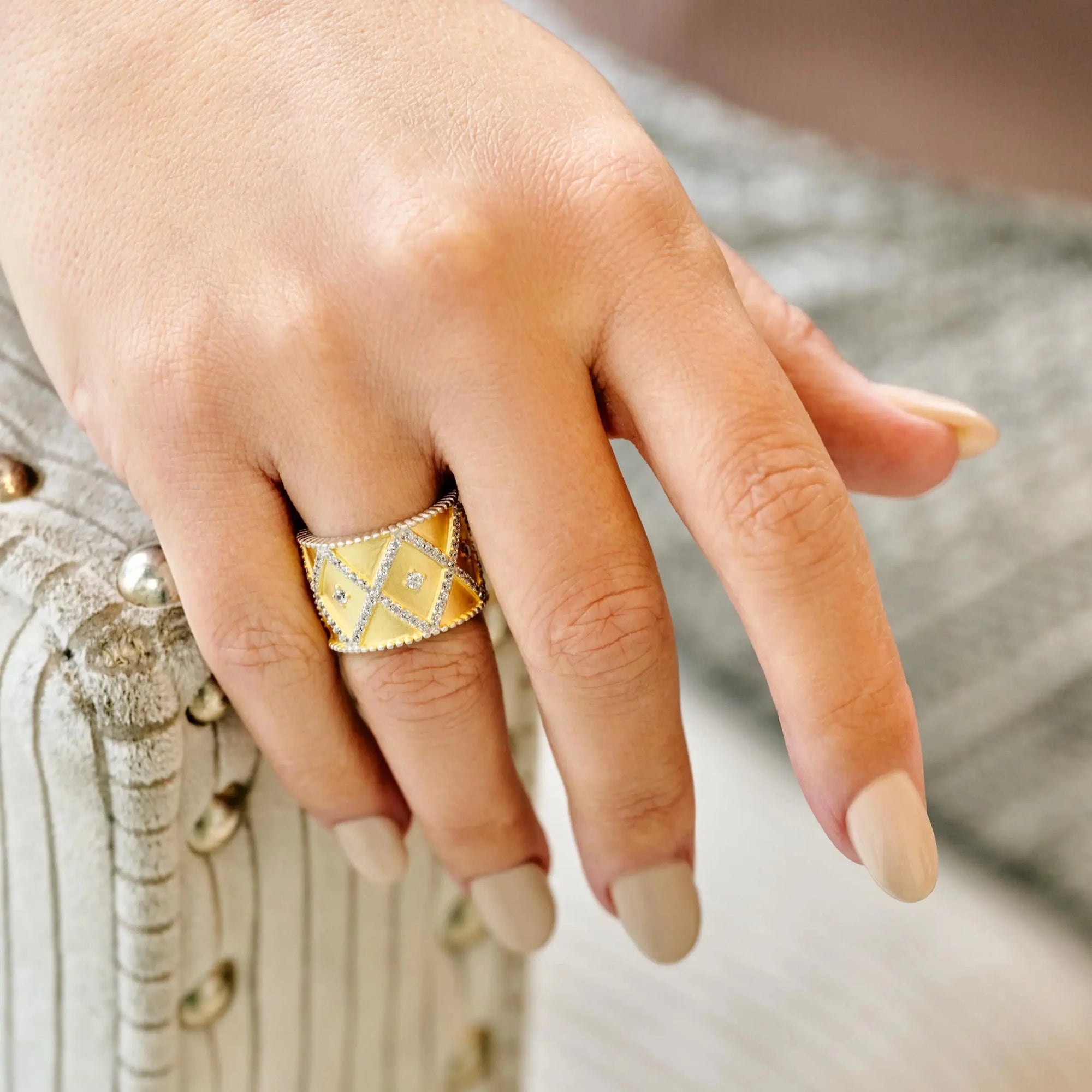 A hand with nude-colored nails rests on a beige textured surface adorned with metallic studs, wearing the All-Time Favorite Cigar Band Ring. The wide gold band, embossed with geometric diamond shapes, captures attention amidst the softly blurred background.