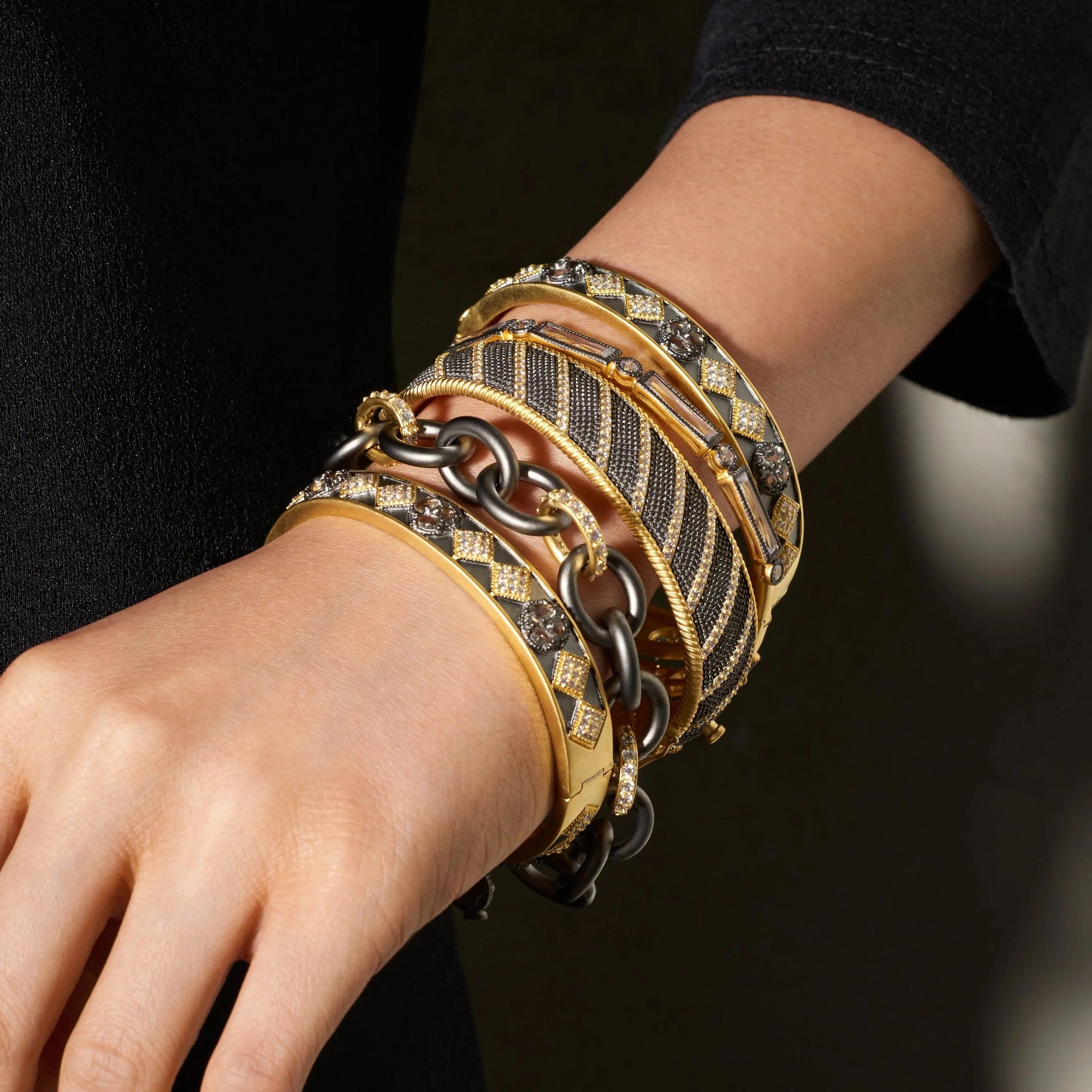 A close-up of a person's wrist adorned with multiple bracelets showcases a luxurious mix, including 14K gold bangles embellished with cubic zirconia-like stones, a black and gold striped design, and the Signature Heavy Link Bracelet featuring a chunky dark chain. The person wears a black sleeve, highlighting the intricate bracelet details.
