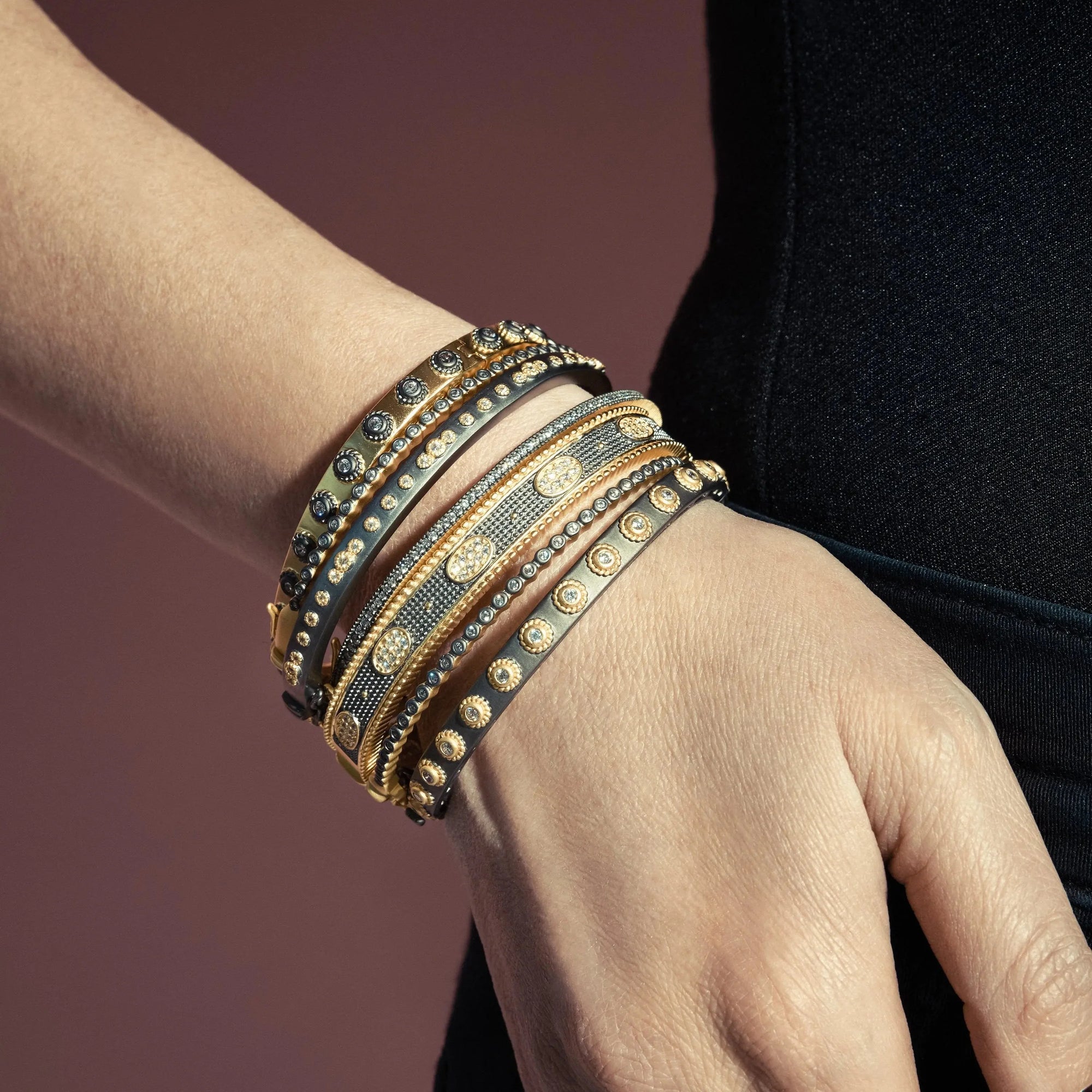 Close-up of a person's wrist adorned with multiple bracelets in stacking styles. The collection features the Studded Eternity Hinge Bangle among a variety of designs incorporating gold, silver, and black elements with dotted patterns. A muted, warm mauve background complements the black garment worn.