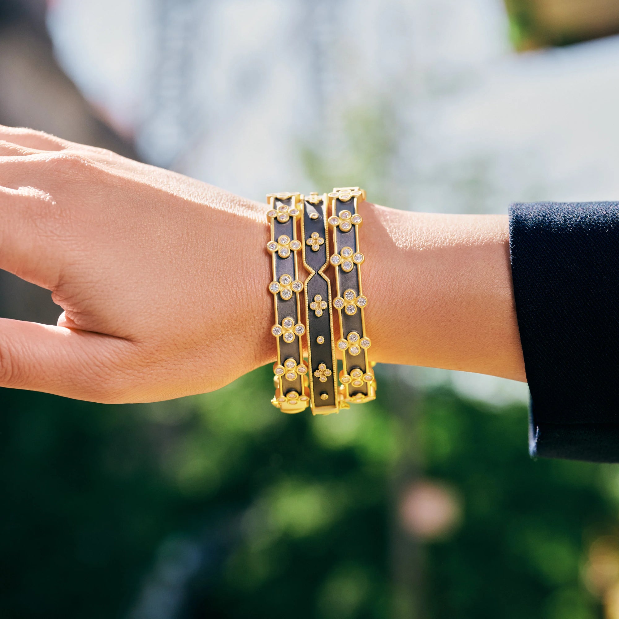 A hand wearing two gold bracelets showcases an intricate design with small, flower-like motifs. One features a black and gold two-tone finish, while the other is the Signature Brooklyn in Bloom Clover Hinge Bangle, dazzling with its entirely golden clover detail. The blurred green foliage in the background adds a natural, elegant setting.