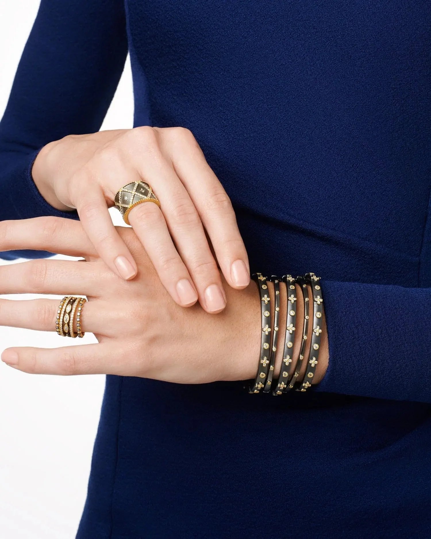 Close-up of a person wearing a dark blue, long-sleeve top. They sport the All-Time Favorite Cigar Band Ring, featuring a large, domed design in gold, on one hand and a multi-band gold-studded bracelet on the wrist. The nails are neatly manicured and the skin tone is light.