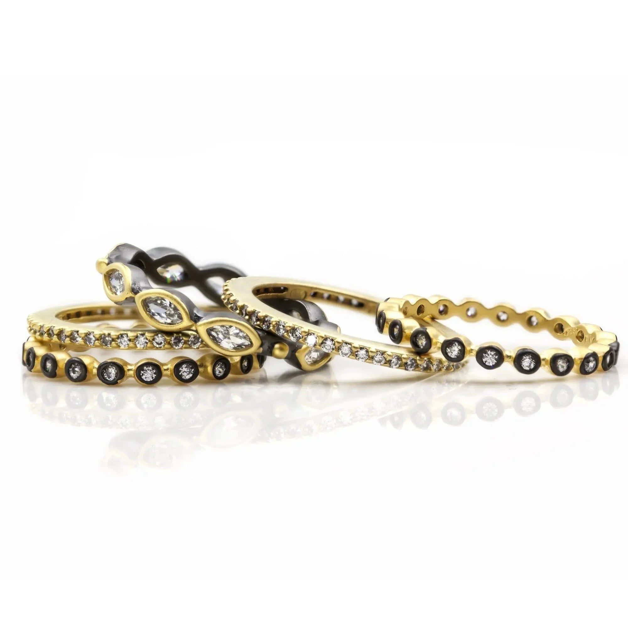 The Signature Marquise Station Ring Stack is a collection of elegant stackable rings showcasing diverse designs, including a gold ring with black inlay and diamonds, two silver bands with marquise-cut diamonds, a thin gold band adorned with small diamonds, and a two-tone gold ring featuring round diamonds and black accents, all beautifully arranged for a stylish display.