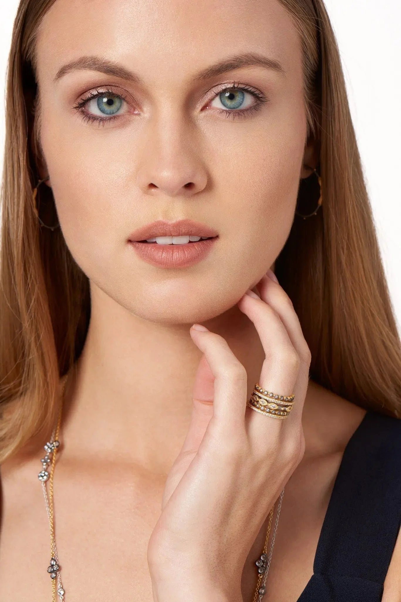 A woman with long, straight hair gazes directly at the camera, her blue eyes and subtle makeup highlighting her elegance. She adorns hoop earrings and a gold necklace, and on her left hand near her chin, she wears the Signature Marquise Station Ring Stack. Her dark outfit features a V-neckline against the plain white background.