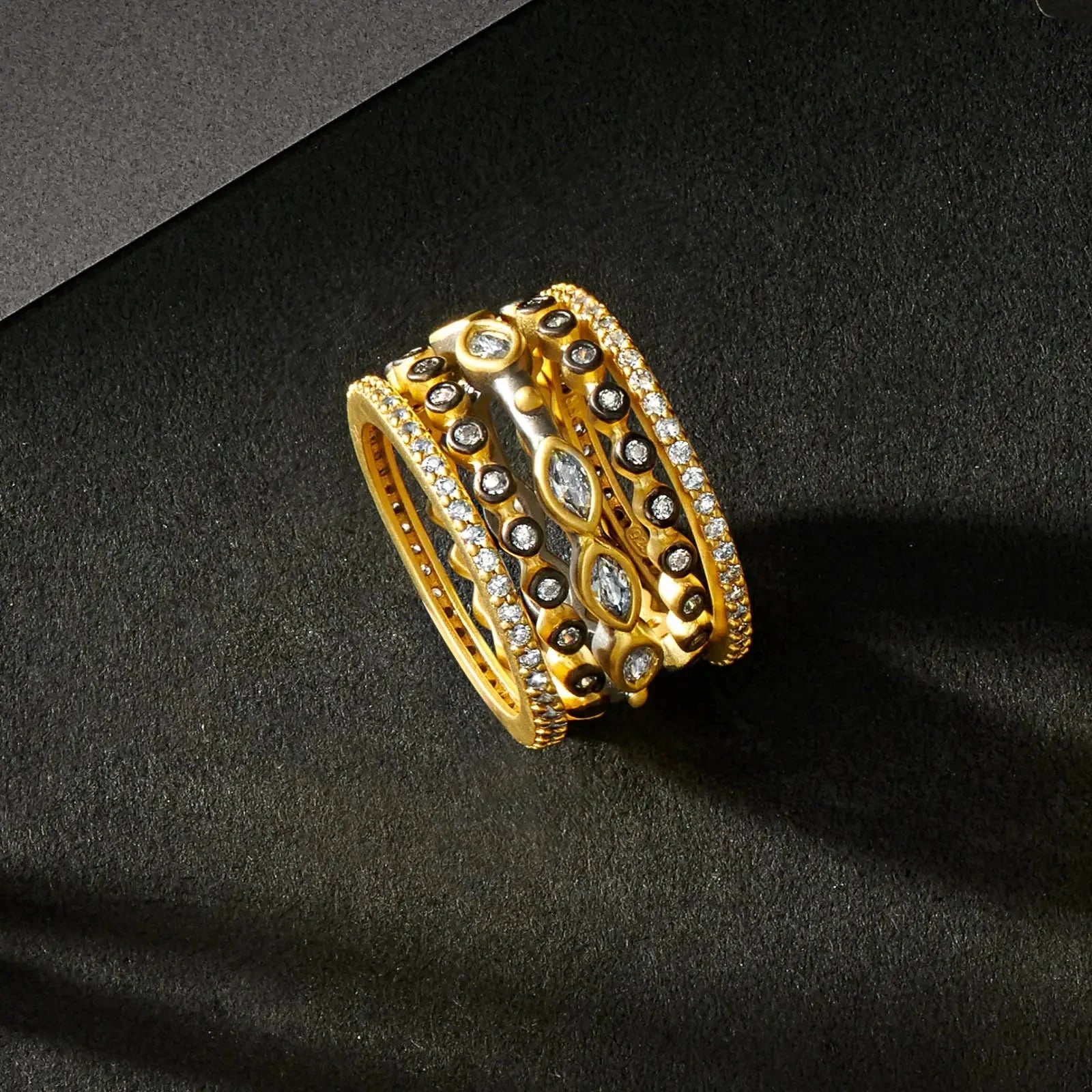 A Signature Marquise Station Ring Stack with exquisite detailing rests on a dark surface. This piece, from a collection of stackable rings, showcases circular and marquise-shaped diamonds arranged in rows and surrounded by bead-like textures. The dark background enhances its shimmer and intricate design with soft shadows.