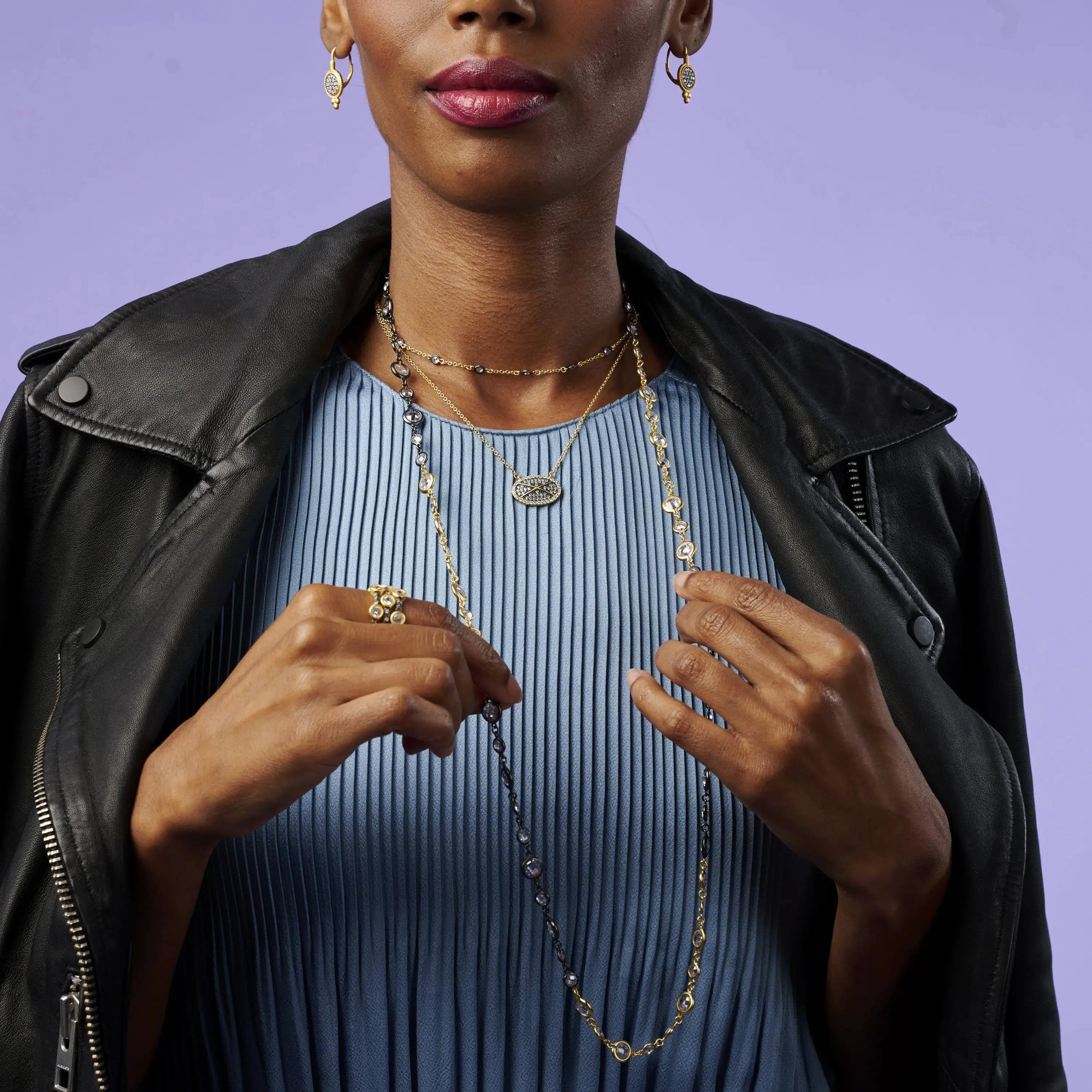 A woman wearing a black leather jacket over a light blue, pleated top stands against a purple background. She accessorizes with gold earrings, layered necklaces, and the Starry Night Ring Set. Her hands hold one of the necklaces while her lips are painted in a deep pink shade.