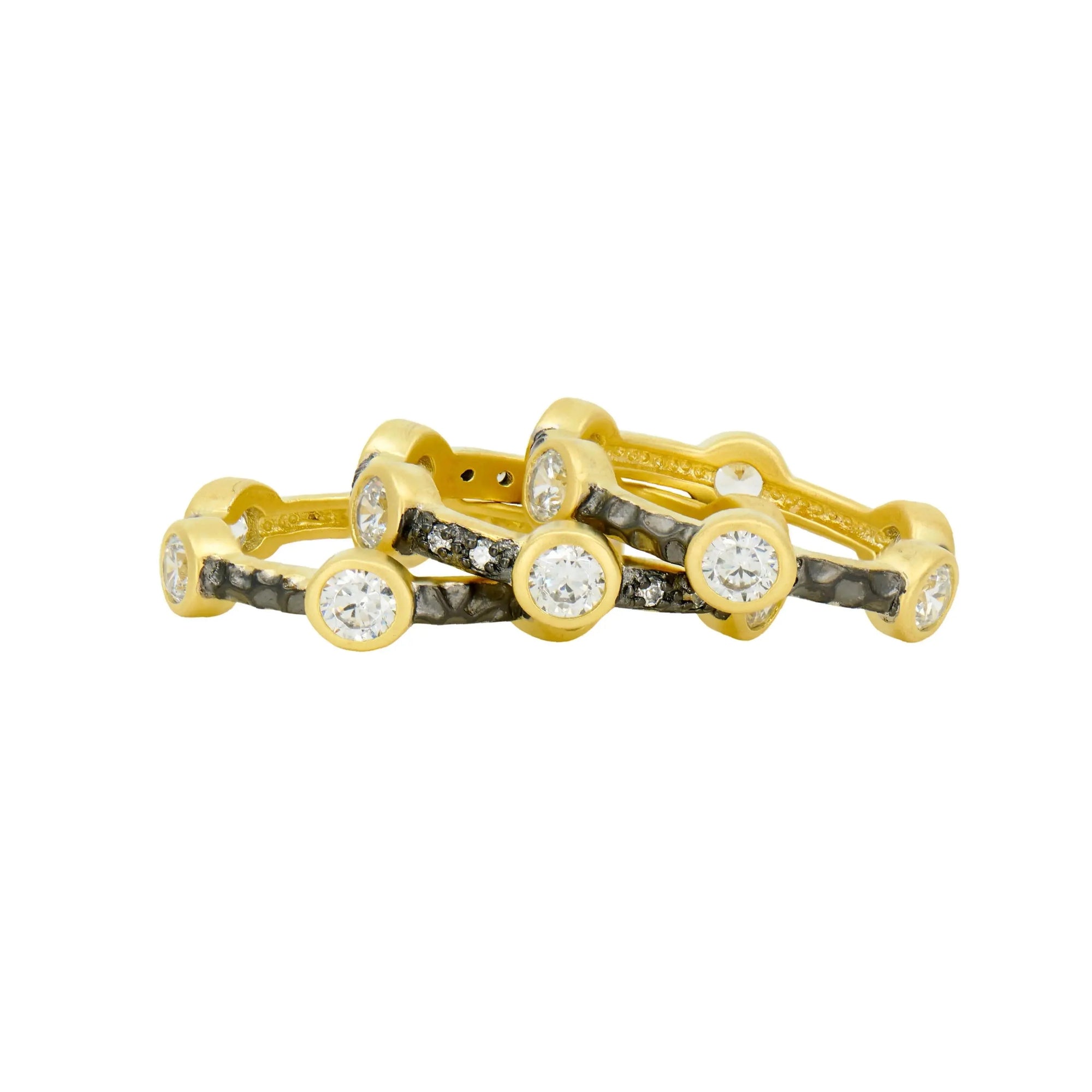 Presenting the Starry Night Ring Set: a trio of entwined gold rings, each adorned with prominent round diamonds elegantly set in bezels. These rings boast a hammered dark rhodium finish and are accentuated with smaller black stones nestled between the larger diamonds, creating a luxurious and intricate look.