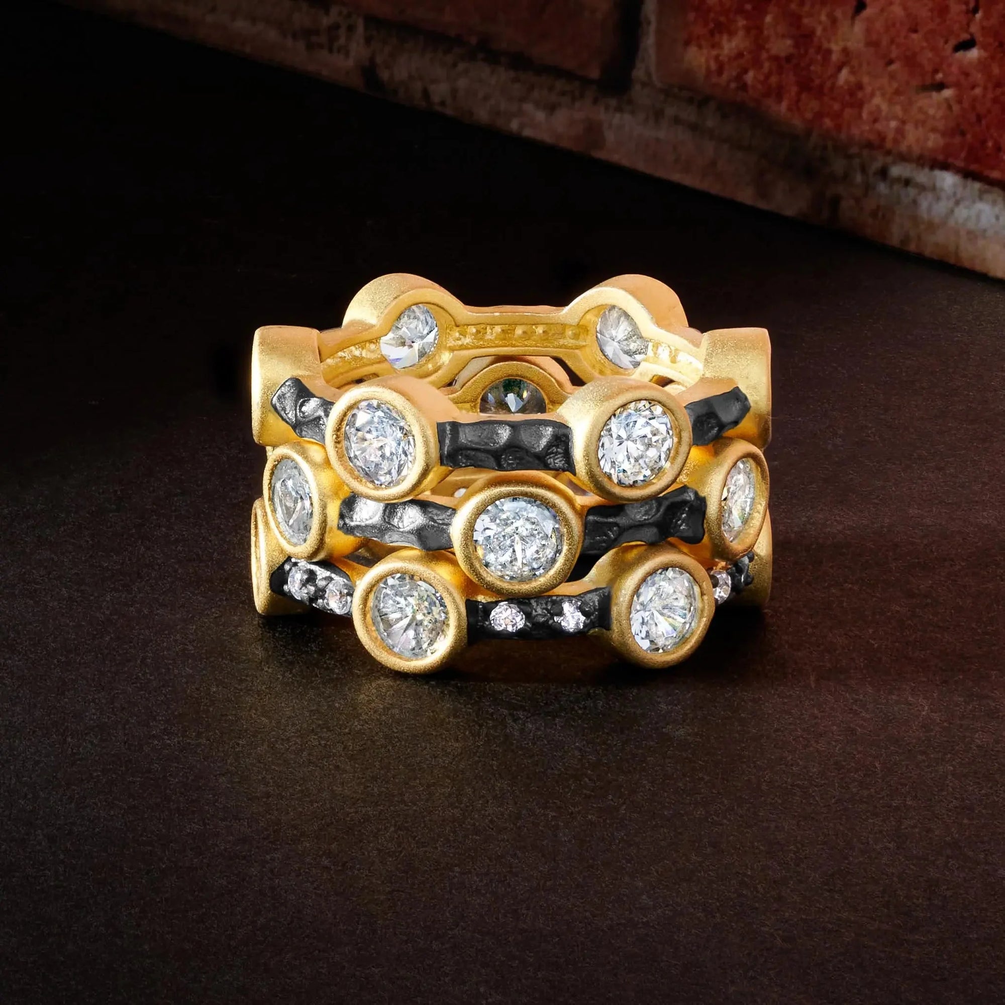 The Starry Night Ring Set features a trio of gold rings embellished with large round diamonds, set against a sleek black surface. Two of the rings showcase a matte finish accentuated by alternating black accents, while the top ring shines with a smooth polish. A blurred red brick wall provides the backdrop for this elegant arrangement.