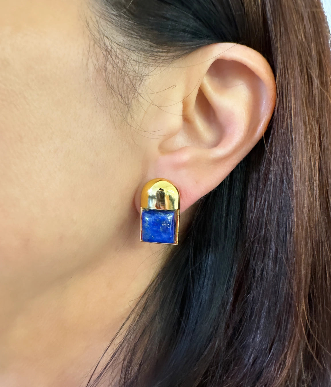 Close-up of an ear showcasing the Baby Cubism Earrings, featuring a blue rectangular stone set in 14 karat gold. This modern design contrasts elegantly with the person's dark brown hair, and its hypoallergenic silicone back ensures comfort for sensitive ears.