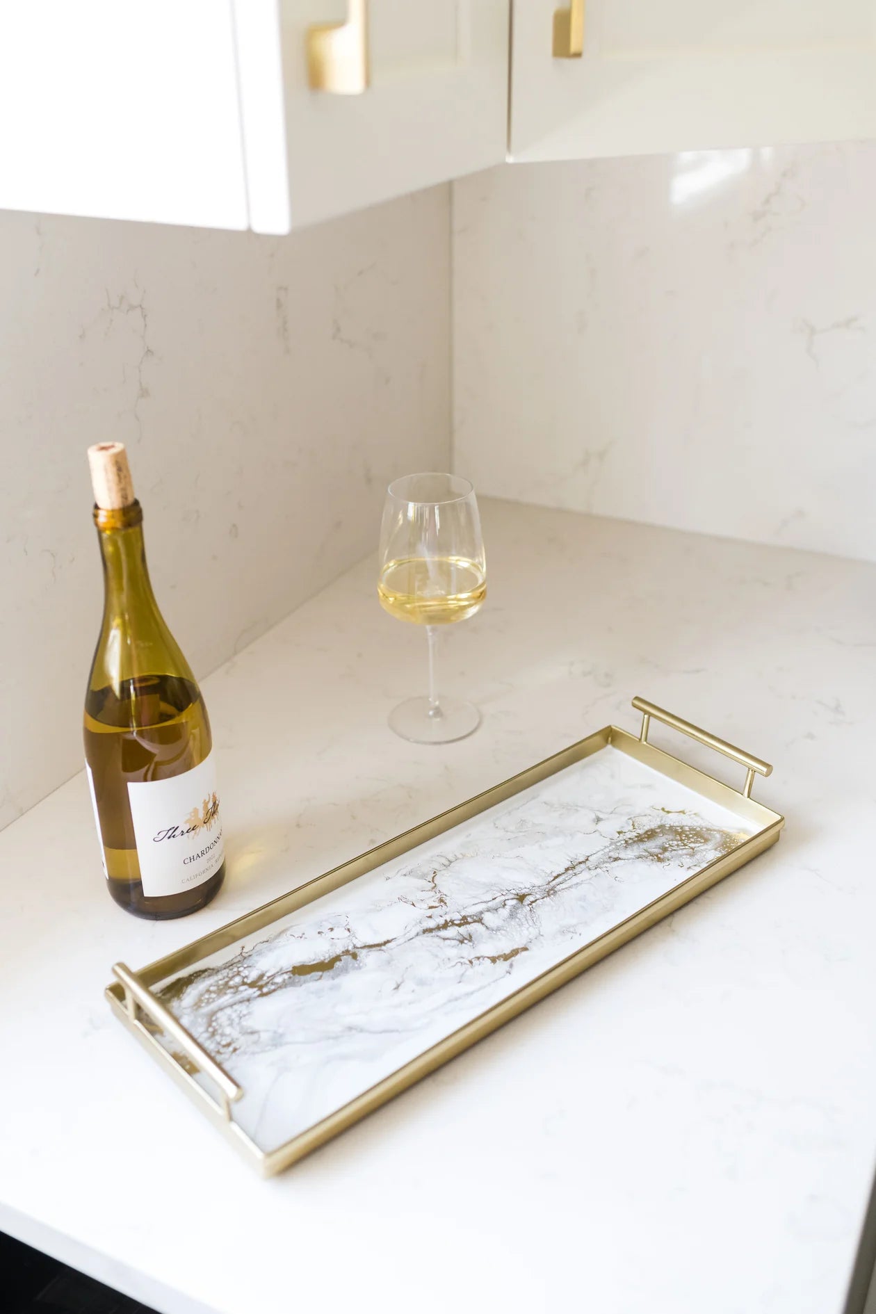 A Resin Gold Rectangle Tray elegantly rests on a white countertop, adorned with matte gold handles. A bottle of Chardonnay and a filled wine glass complement the scene, embodying elegance in this modern kitchen with white cabinets. The handmade artisan touch enhances its charm, making the setting both minimalistic and sophisticated.