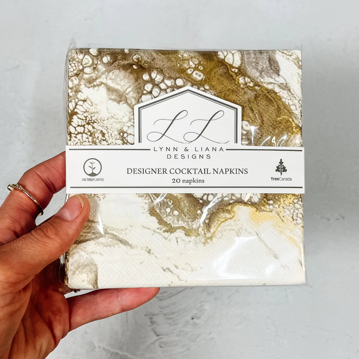 A hand holds a pack of Lynn & Liana Designs Cocktail Napkins, showcasing an abstract marbled pattern in gold, white, and brown. The packaging indicates it contains 20 premium 3-ply napkins and mentions that partial proceeds support Tree Canada.
