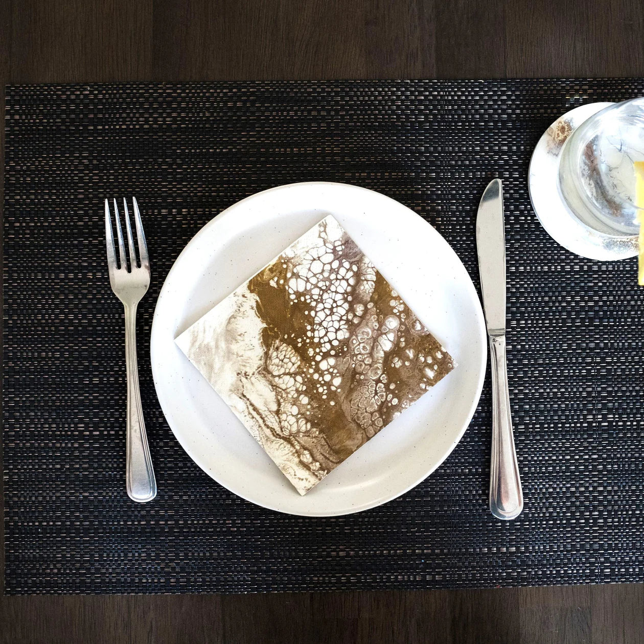 A white plate, accompanied by premium Cocktail Napkins, is set on a dark woven placemat. To the left of the plate is a fork, while a knife rests on the right. In the upper right corner, there's a small glass of water with lemon alongside a butter dish placed on a matching saucer. The table surface is dark wood.