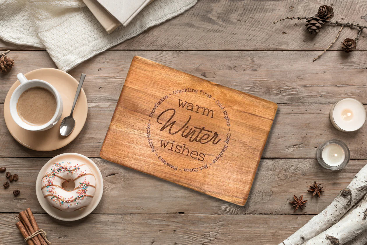 An Engraved Board embellished with "Warm Winter Wishes" and encircled by festive words is positioned on a rustic table. Alongside, there's a cup of coffee, a donut topped with sprinkles, candles, pinecones, and star anise. A white textured cloth and sheets of parchment enhance the cozy winter ambiance.