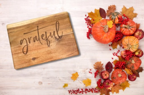 On the left is the Engraved Board, a wooden acacia cheese board with "grateful" elegantly written in cursive. On the right, a holiday charcuterie board showcases autumnal elements such as pumpkins, apples, pinecones, cranberries, and an array of colorful leaves on a light wood surface.