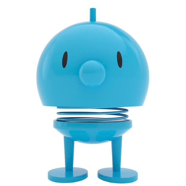 Introducing the Hoptimist Bumble, a delightful toy figure in blue featuring a round head and body, wide-set black oval eyes, and a small nose. The spring connecting its head and body adds a playful touch to your home decor. It stands upright on two flat, round feet, perfectly capturing the cheerful essence of Danish design history.