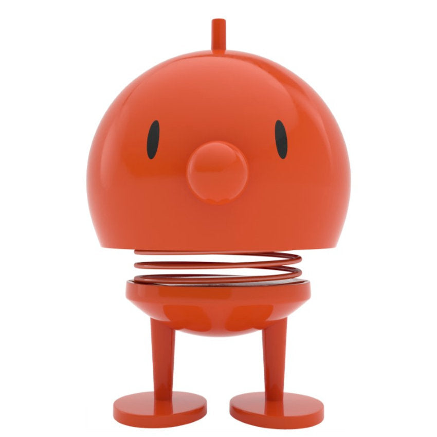 The Hoptimist Bumble, steeped in Danish design heritage, is a small, round orange figure with an uncomplicated, cartoon-like face that boasts black oval eyes and a round nose. Its spring-loaded body delivers a whimsical bounce as it rests on two short legs with flat circular feet—an enchanting addition to any home decor.