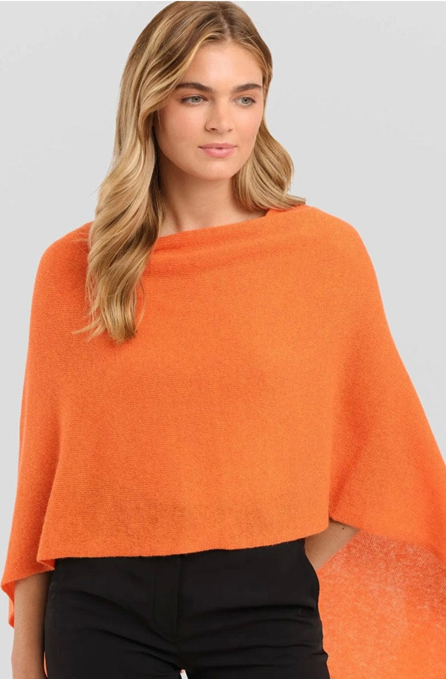 A woman with long blonde hair is adorned in a vivid orange Cashmere Poncho, elegantly draped over her black outfit. This one-size dress topper gently cascades over her shoulders as she poses against a light gray backdrop, glancing slightly to her left with a serene demeanor.