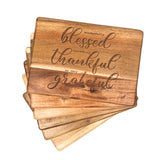 A set of the Engraved Board coasters, featuring the words "blessed," "thankful," and "grateful" in cursive, pairs beautifully with an Acacia cheese board. These light brown coasters, displaying natural wood grain patterns, are elegantly arranged against a crisp white background.