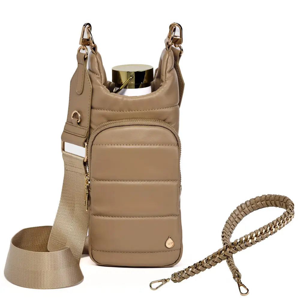 The Vegan Leather HydroBag® with Strap Bundle is highlighted, featuring a tan, quilted crossbody design with an adjustable beige strap and gold detailing. Made from vegan leather, it includes an outer zippered pocket and a detachable braided strap. A distinctive gold bottle cap suggests the presence of a water bottle inside. Accompanying the bag is a matching lanyard displayed elegantly alongside it.