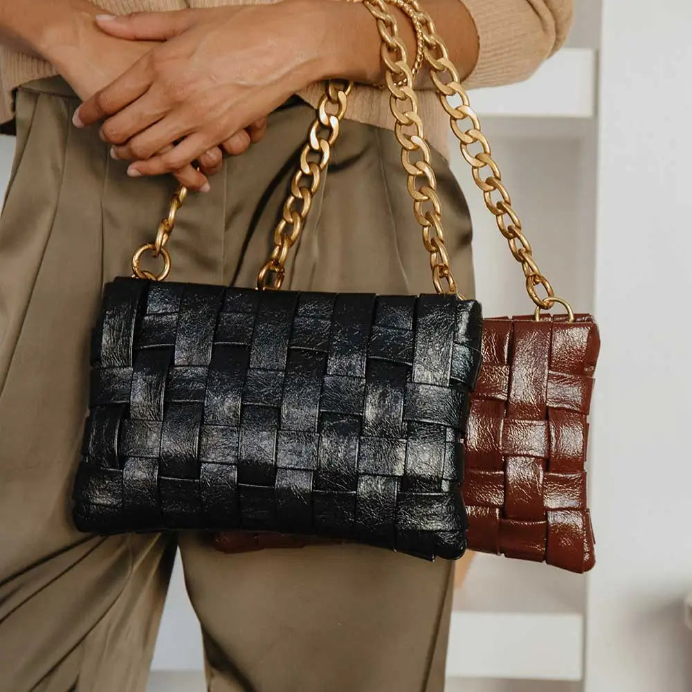 A person holds two Mimie Recycled Vegan Crossbody Bags with thick gold-tone hardware chain straps. One is black, and the other is brown. They're wearing beige pants and a sweater against a blurred background of a white wall and shelves.