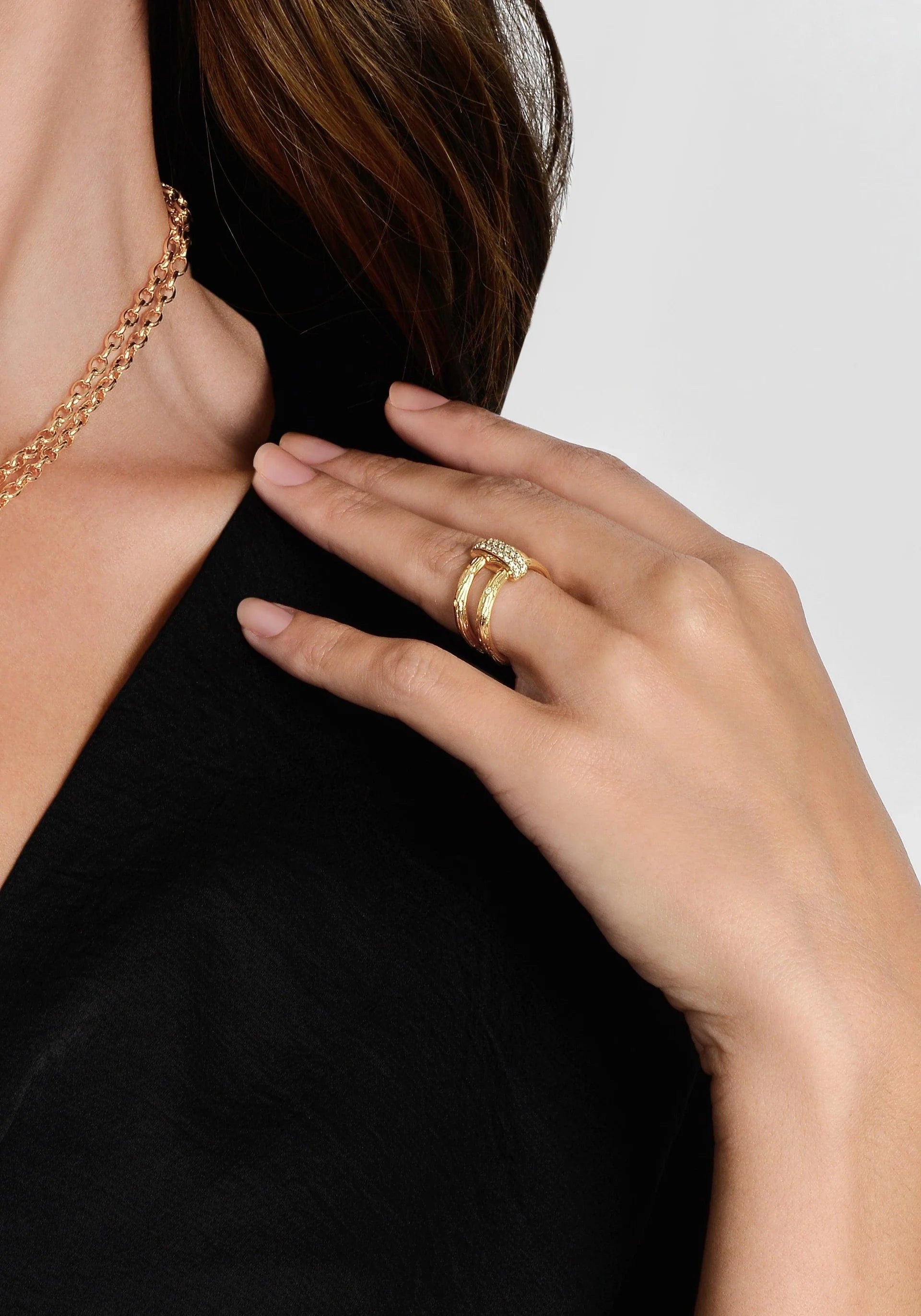 A person in a black top is captured from the neck to the hand, showcasing a layered gold necklace and an Enchanted Forest Chain Double Ring on their ring finger. The double rings are embellished with small, sparkling crystals. The person's hair is partially visible, flowing over their shoulder against a plain, light-colored background.