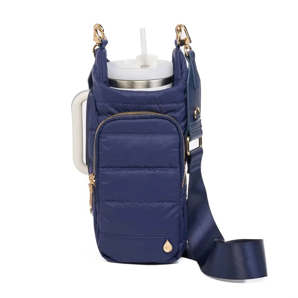 The HydroBag Handle is a navy blue quilted tumbler holder with a gold zipper and hardware, featuring an adjustable strap. It securely holds a stainless steel tumbler with a white lid and straw, and the front pocket provides extra storage for mess-free hydration on the go.