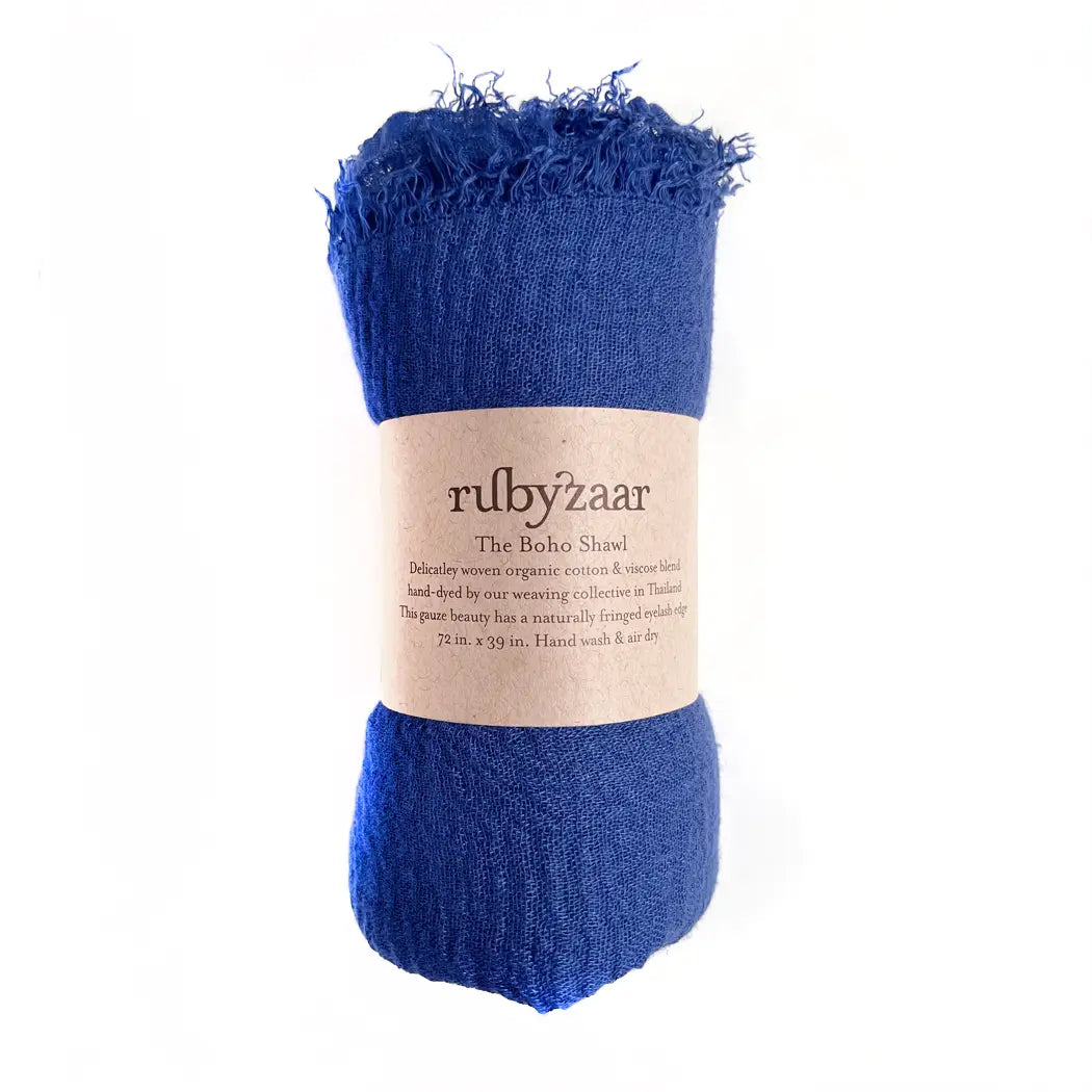 A rolled-up blue shawl with a slightly frayed edge is wrapped in a brown paper label reading, "rubyzaar Boho Scarf." It describes the item as a delicately woven bohemian-style scarf made from organic cotton, hand-dyed in Thailand, with dimensions of 72 x 35 inches.