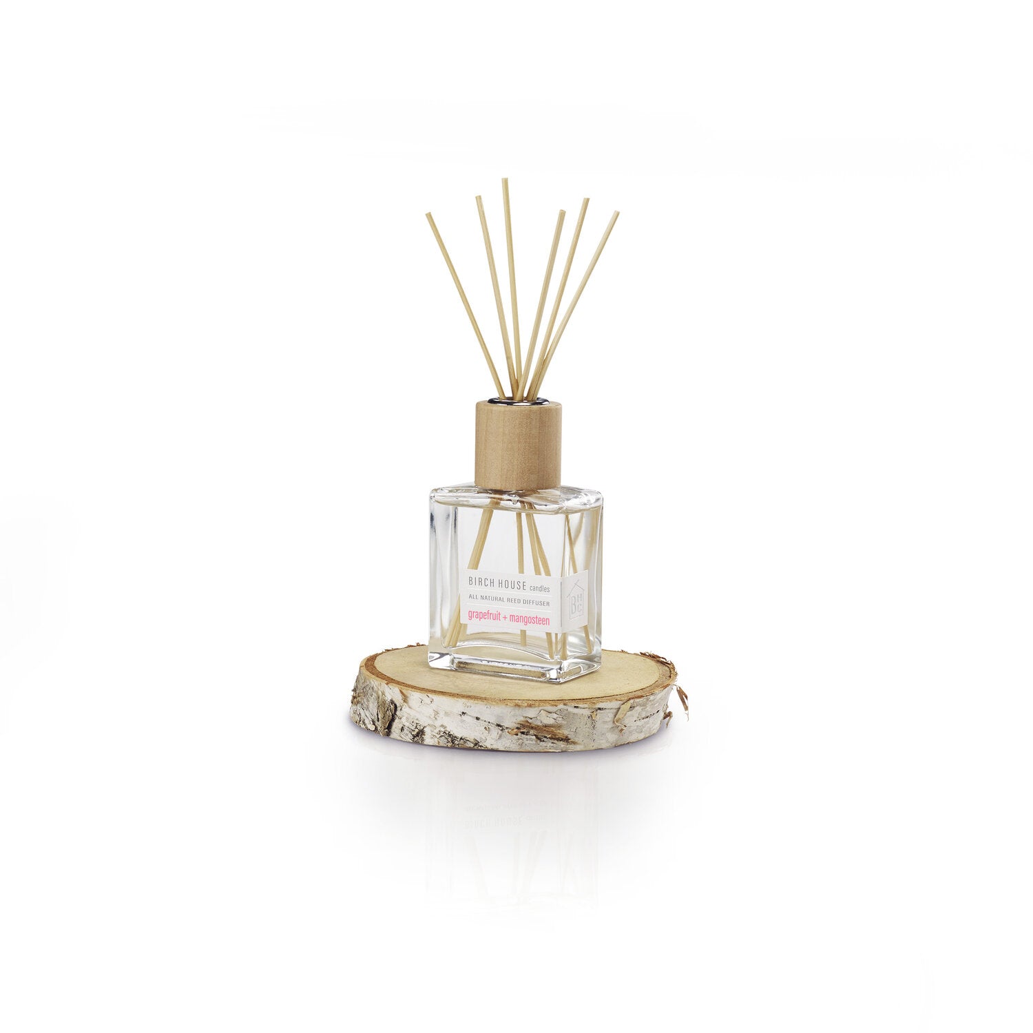A minimalist square glass bottle with a wooden cap holds reed sticks, infused with essential oils for the Sweet Grapefruit Diffuser fragrance. This "Birch Bliss" scent hints at sweet grapefruit and sits on a circular birch wood slice against a plain white backdrop.