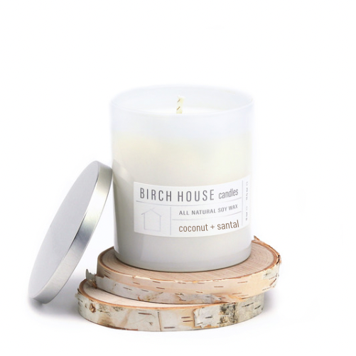 The Coconut + Santal Candle, made from all-natural soy wax and presented in frosted glass with a silver lid, features a soothing scent. It's labeled "BIRCH HOUSE candles" and rests on birchwood slices, embodying a minimalist aesthetic reminiscent of the French Riviera.