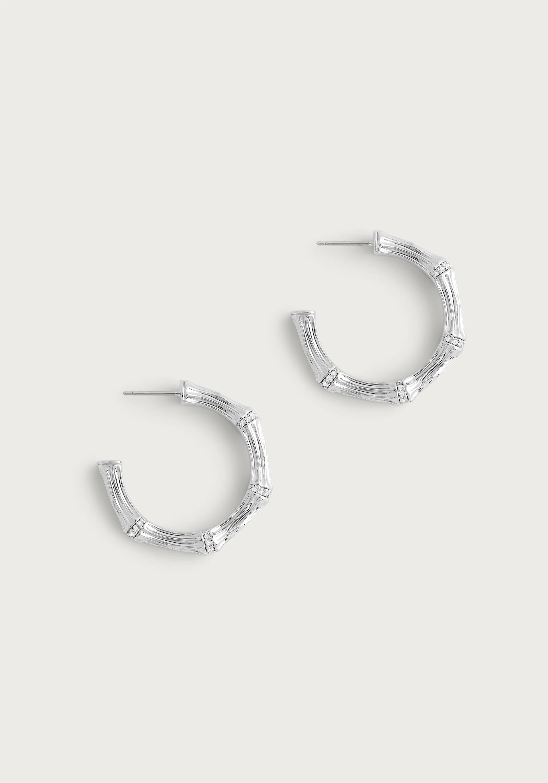 The Bamboo Medium Hoop Earrings - Silver by Anabel Aram are crafted from sterling silver plated brass, featuring a bamboo-like texture and segmented sections for a natural, wood-inspired look against a light, neutral background.